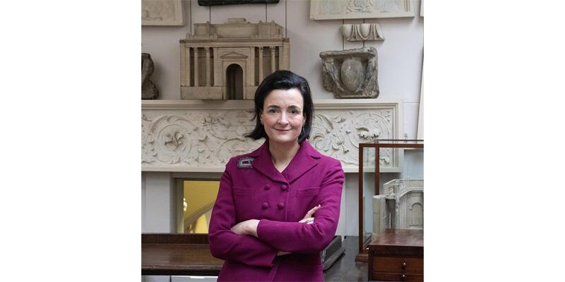 Ursola Rimbotti, wearing a pink suit, stands with her arms folded in the Drawing Office.